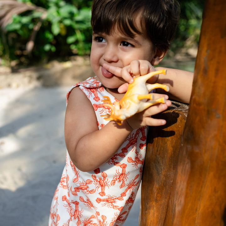 Child holding a piece of fruit behind wooden bars in an outdoor setting. Wearing cream handmade onesie with red lobster on it.