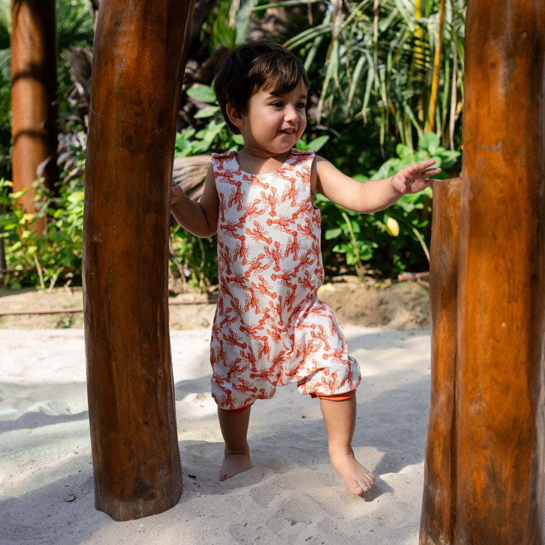 Child in a floral romper standing between two wooden posts in a natural setting. wearing cream handmade onesie with red lobster on it.