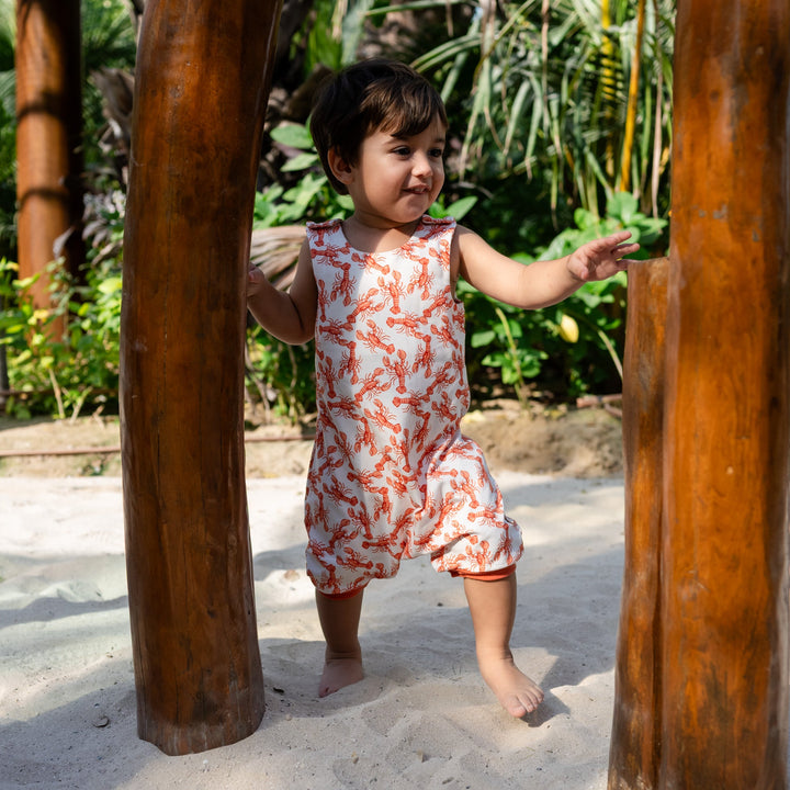 Child in a floral romper standing between two wooden posts in a natural setting. wearing cream handmade onesie with red lobster on it.
