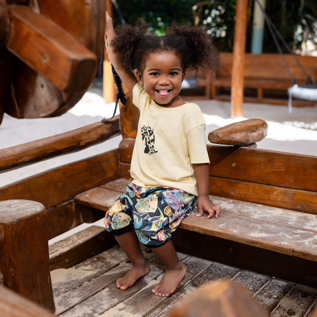 Child sitting on a wooden swing set with a smile. Wearing a handmade organic set shorts and shirt with color full fishes and yellow t shirt