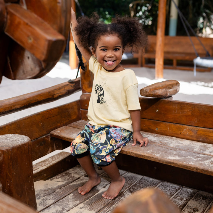 Child sitting on a wooden swing set with a smile. Wearing a handmade organic set shorts and shirt with color full fishes and yellow t shirt