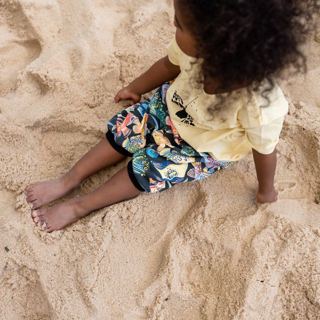 Child playing in sand with colorful shorts on a beach. Wearing a handmade organic set shorts and shirt with color full fishes and yellow t shirt