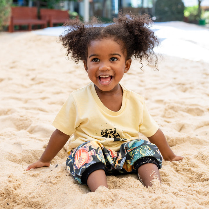 Child sitting in a sand pit at a playground. Wearing a handmade organic set shorts and shirt with color full fishes and yellow t shirt