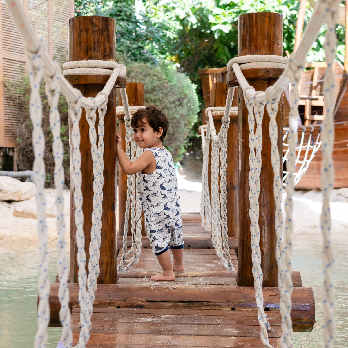Child standing on a wooden bridge over water with rope railing, surrounded by greenery. He is wearing a white handmade onesie with blue sharks on it