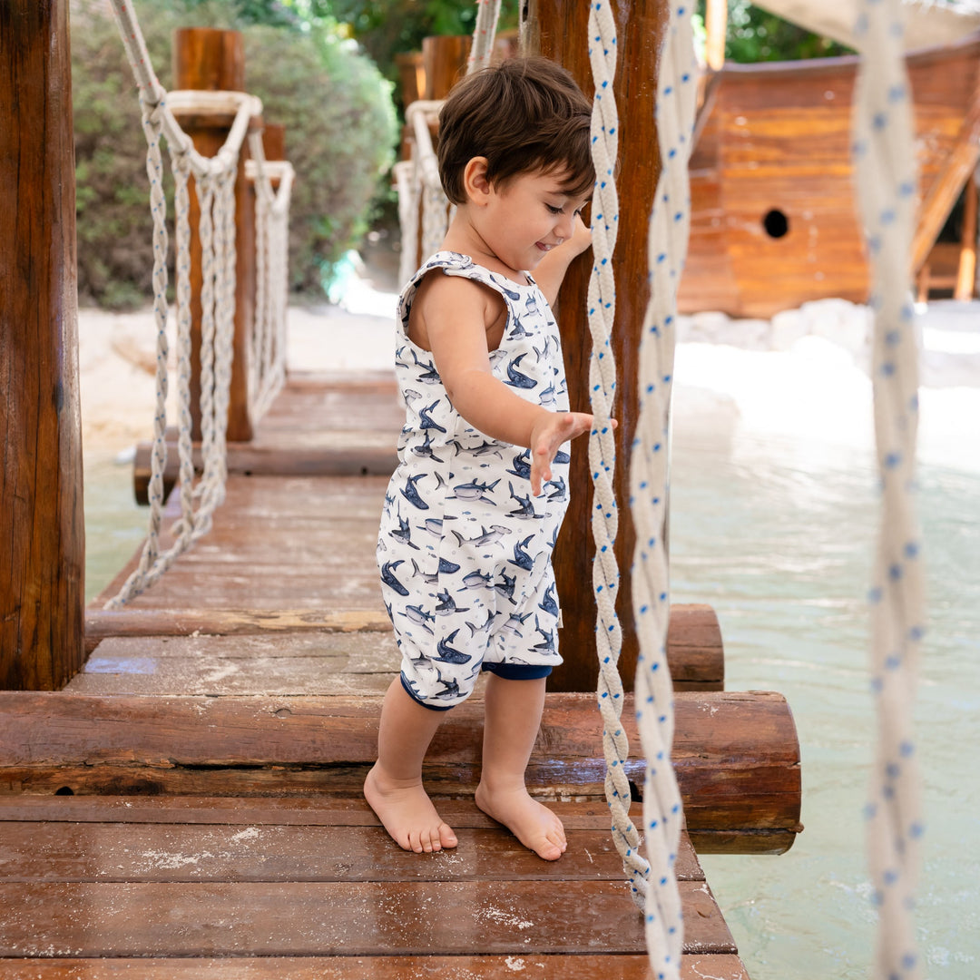 Child standing on a wooden swing set with a playground in the background. He is wearing a white handmade onesie with blue sharks on it