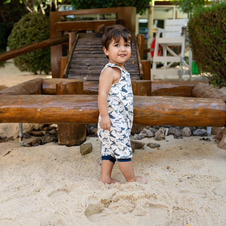 Child standing on a wooden seesaw in a playground. He is wearing a white handmade onesie with blue sharks on it