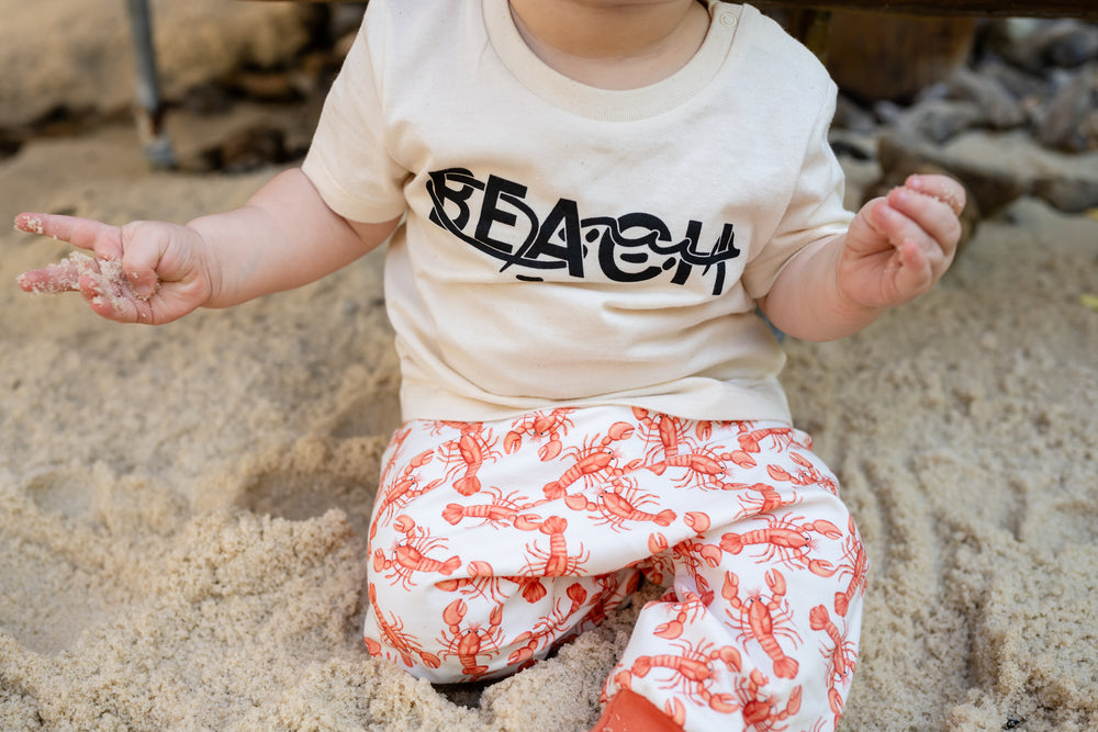 Baby sitting in sand wearing a 'Beach Habibi'T- shirt and harem pants with red lobster on them