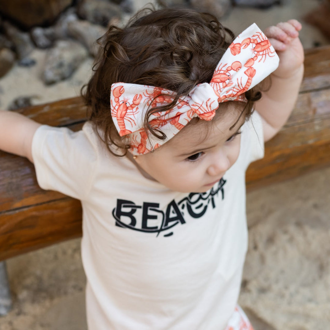 Child standing on a sandy surface with a wooden structure, wearing a white shirt with text and floral shorts. wearing a handmade headband with red lobster on it