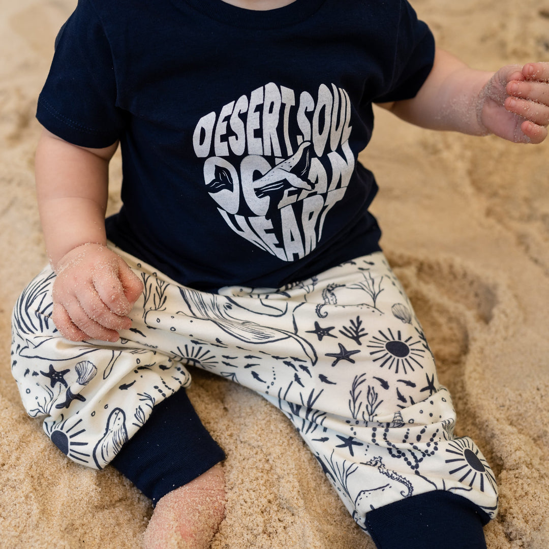 Baby sitting on sand wearing a navy blue shirt with white text and patterned pants.