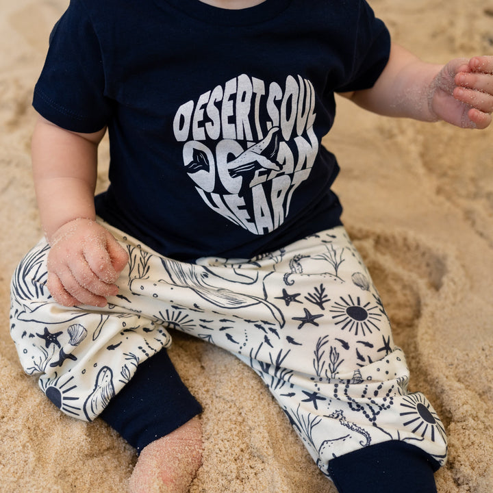 Baby sitting on sand wearing a navy blue shirt with white text and patterned pants.