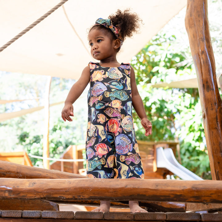 Young girl in a wearing colorful fishes on a black romper standing on a wooden structure with greenery in the background