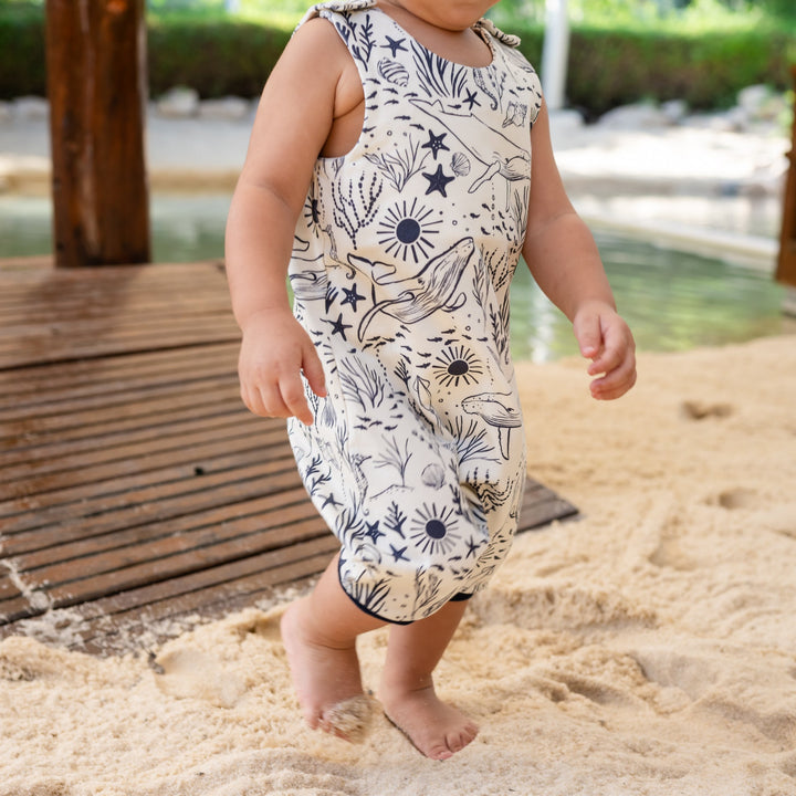 Child in a patterned romper standing on sand with a wooden structure and water in the background