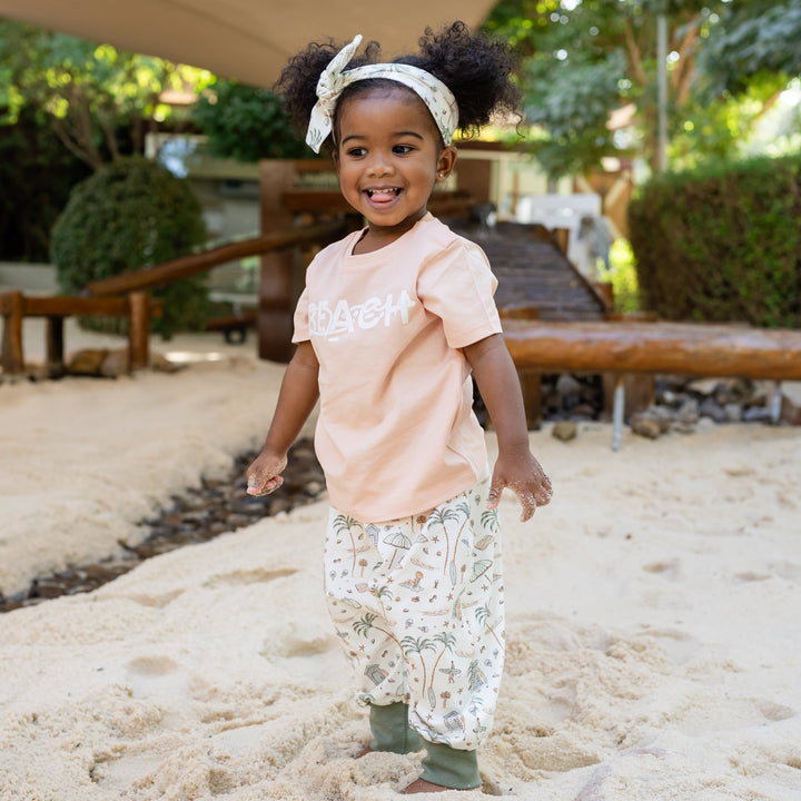 Child standing on a sandy surface with greenery in the background