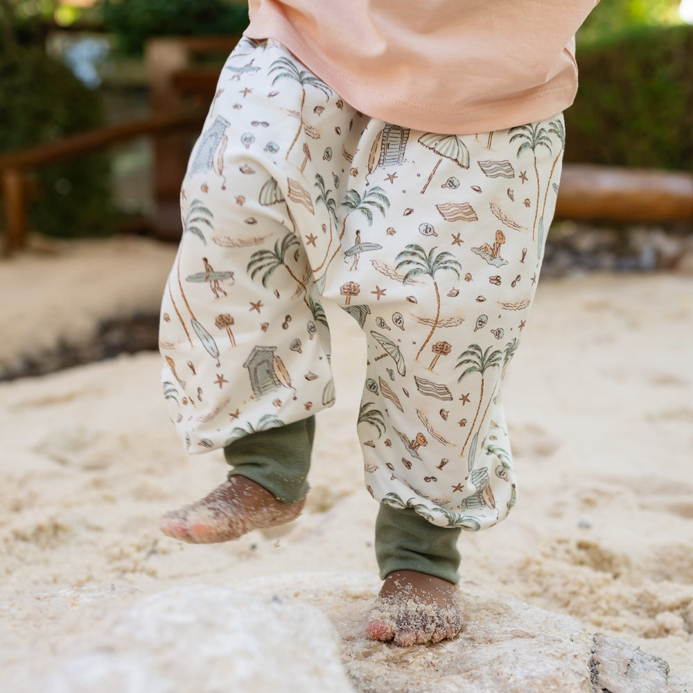 Child wearing patterned pants standing on a sandy surface with greenery in the background