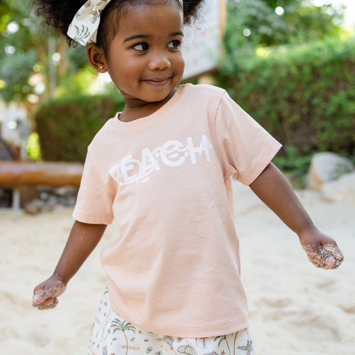 Child wearing a light pink t-shirt with text, standing in a sandy area with greenery in the background.