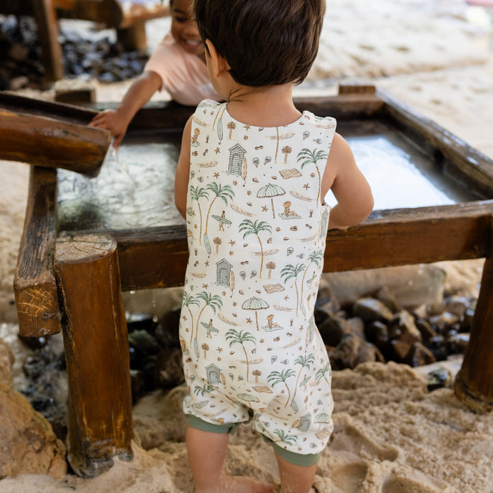 Child in a sand pit wearing a patterned romper, standing near wooden benches.