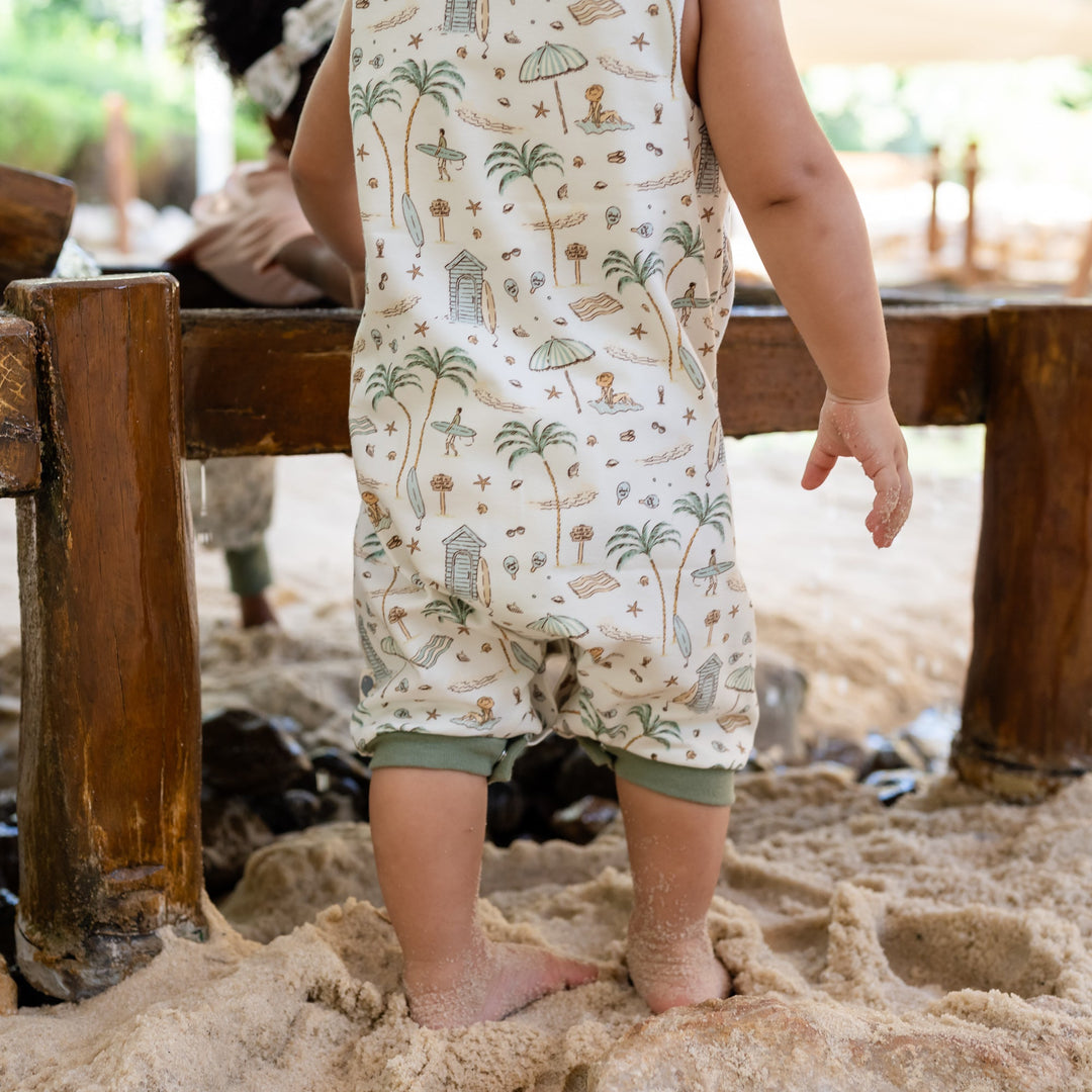 Child in a patterned romper standing in sand near wooden structures
