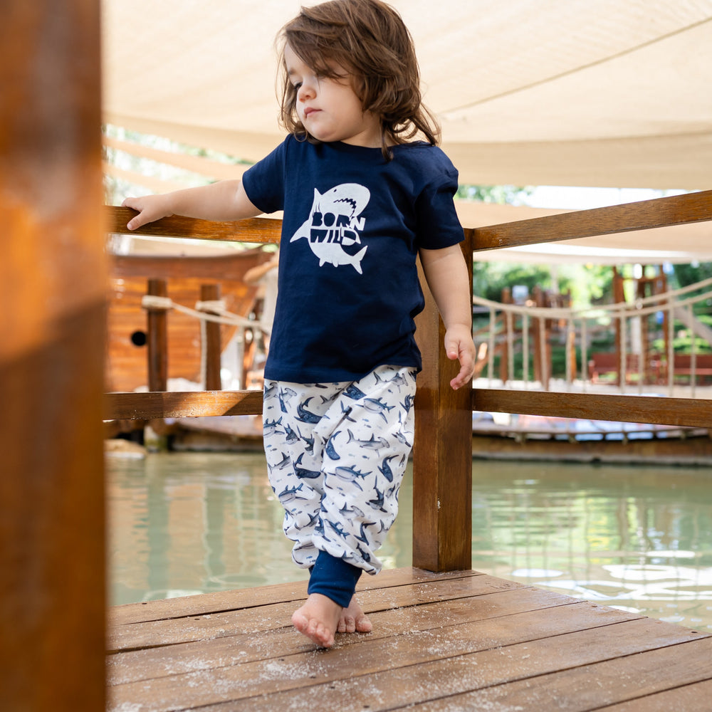 Child standing on a wooden deck by a pool, wearing a navy blue shirt with a white graphic and light-colored pants.